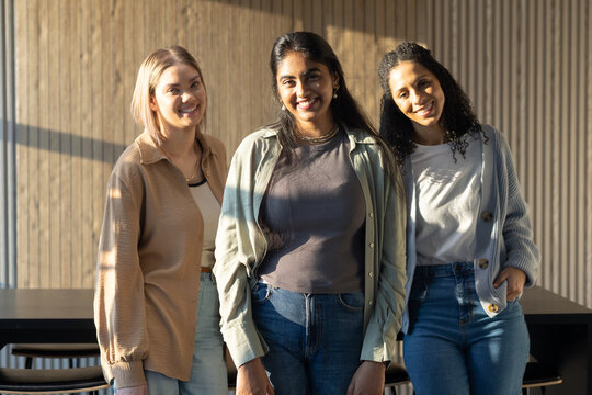 Diverse female friends standing in warmly lit coworking corner by wooden slat wall, long table