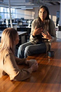 Diverse female coworkers talking and gesturing at open office large wooden table with ceramic mugs