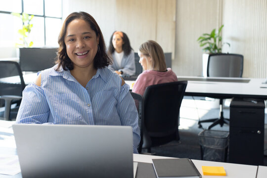 African American woman 30s blue blouse smiling, typing at office desk with laptop and sticky pad