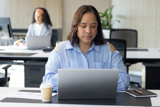 Adult African woman working on laptop at office wearing striped blouse with hoops, coffee cup