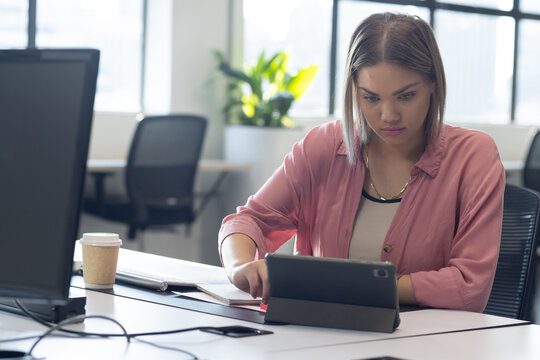 Woman wearing pink shirt and gold chain using tablet in folio case at office desk, notebook