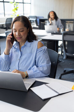African American female coworkers talking on phones, using laptops at open-plan office desks