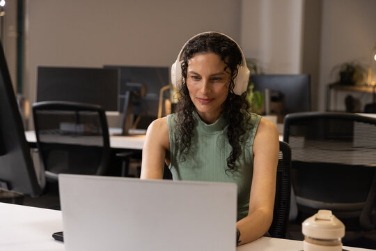African American woman working at laptop in open office wearing green tank top, beige headset