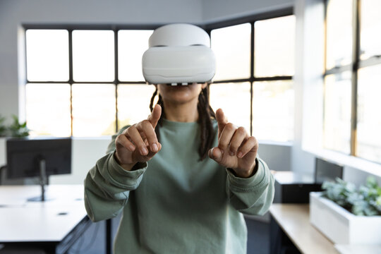Nonbinary person wearing white VR headset, extending fingertips in office with left monitor