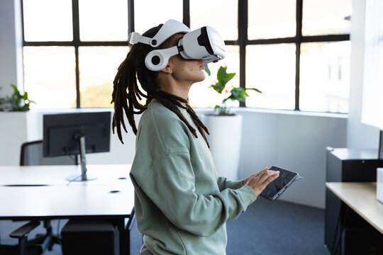 African American nonbinary person wearing sweater VR headset using tablet by work window