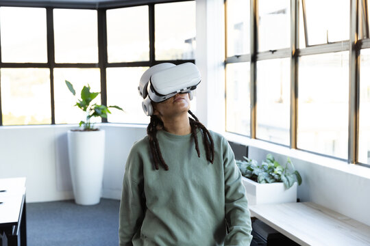 Non-binary adult standing wearing white VR headset and green crewneck by grid windows with planter