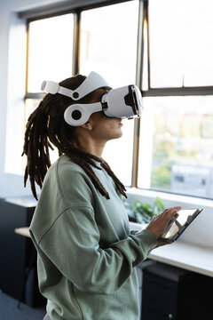 Non-binary African person in green sweater VR headset with tablet testing VR by paned office window