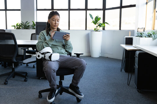 Non-binary African American person sitting in open-plan office holding tablet with white VR headset