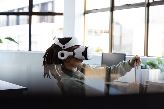 Non-binary person with dreads sitting in swivel chair, wearing VR headset and holding laptop