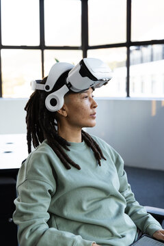 Nonbinary African American wearing VR-headset sage-top seated in office using tablet by grid window