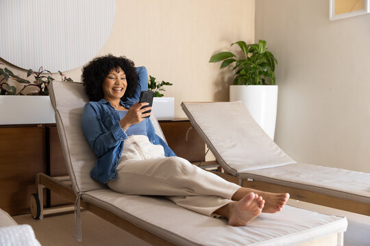 African American woman reclining on chaise lounge at spa lounge, checking smartphone barefoot