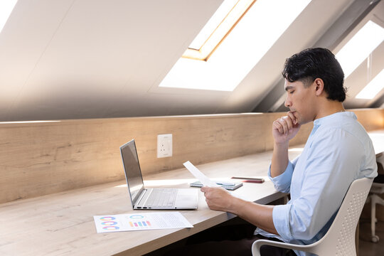 Man in button down shirt sitting at loft desk reviewing charts on laptop, smartphone, copy space