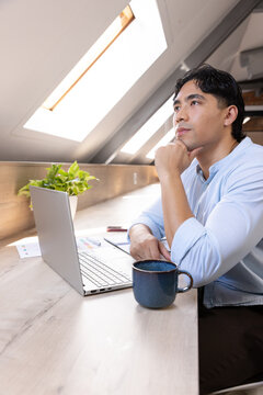 Asian man sitting in loft wearing blue shirt, gazing to skylights, laptop, coffee mug, copy space