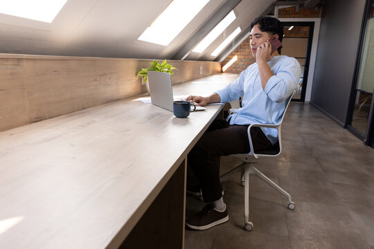 Asian man sitting at loft office bench under skylights, holding phone, typing on laptop, copy space