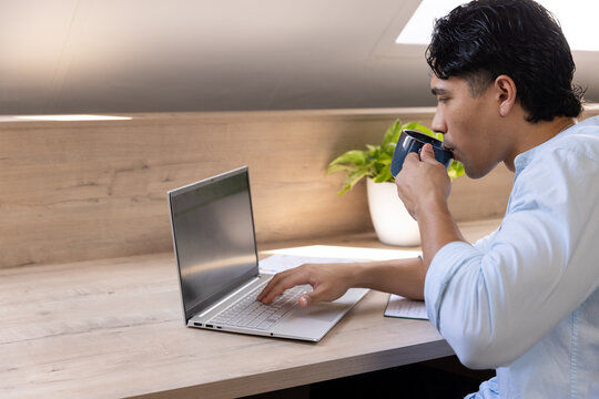 Asian man sitting at desk under skylight sipping blue mug, typing on laptop, copy space