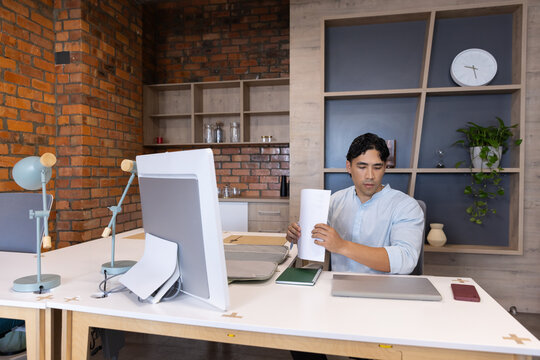 Adult Asian man sitting at white worktable in office examining papers near monitor, copy space