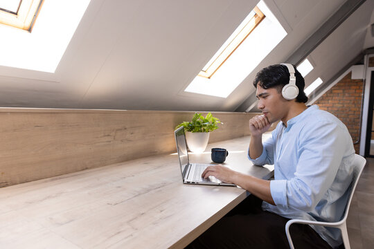 Asian man working on laptop at loft counter wearing headphones and holding mug, copy space