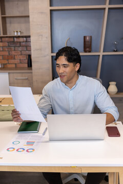 Asian man wearing light blue shirt reviewing documents and working on silver laptop at desk