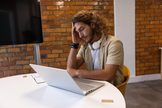 Male sitting at round meeting table, resting head, looking at silver laptop with headphones