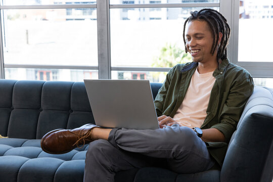 African American man typing and smiling on laptop while on blue tufted sofa by windows