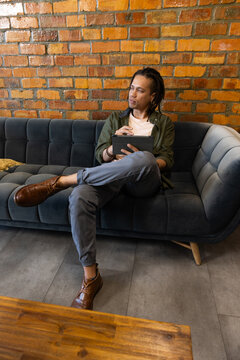 African American man sitting on tufted sofa against brick wall, holding tablet and stylus