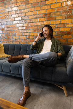 African American man holding smartphone, reclining on gray sofa with mustard cushion and boots