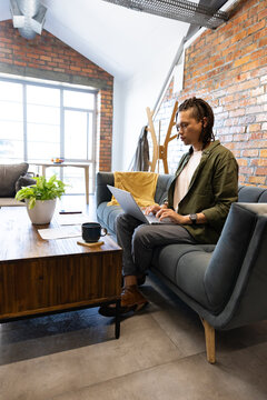 African American adult man working on laptop on sofa with coffee mug, papers, smartphone
