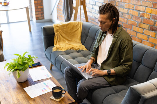 Adult African male, green shirt, sitting in loft on gray sofa typing on laptop, charts, mug