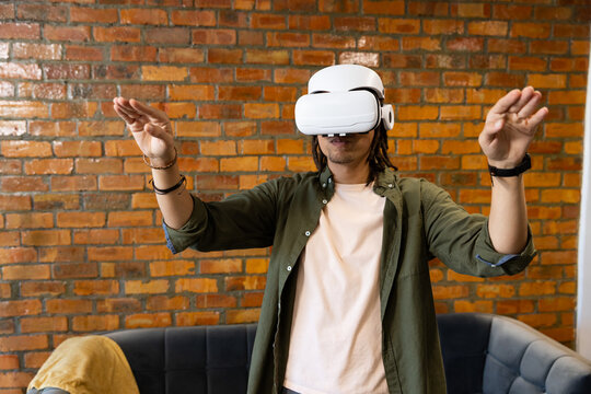 African American man standing wearing VR headset extending arms at sofa, mustard throw, brick wall