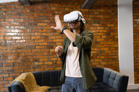 African adult male wearing white VR headset, olive overshirt, standing in loft lounge by brick wall