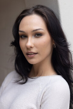 Woman posing near pale wall indoors, wearing light knit top subtle makeup, hoop earrings