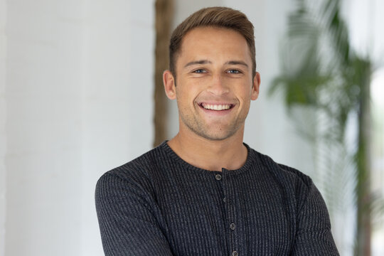 Adult male facing camera in bright room smiling with crossed arms wearing dark henley, plant behind