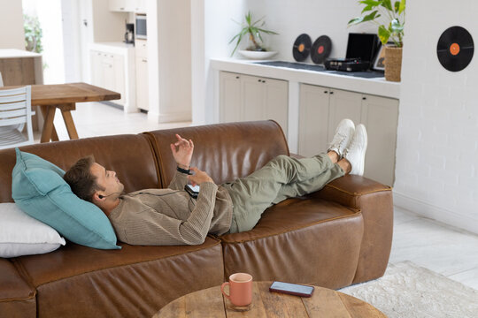 Man reclining on leather sofa in living room, wearing earphones, smartphone on table
