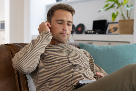 Man reclining on sofa in beige sweater listening with wired earbud and portable player