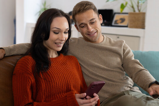 Couple sitting on couch at home, woman wearing rust sweater holding smartphone while man leaning in