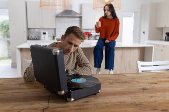 Couple leaning over portable turntable on wooden table in modern kitchen, woman holding mug