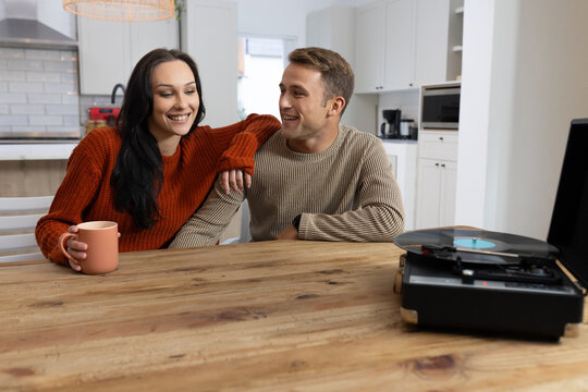 Couple sitting at wood table in kitchen, woman in rust sweater holding mug, record player