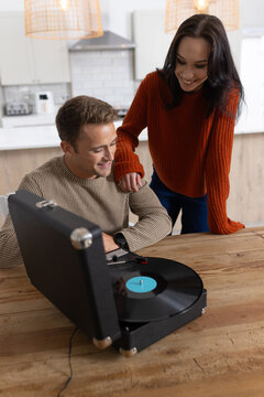 Couple leaning toward suitcase turntable at wooden table in kitchen, handling black vinyl