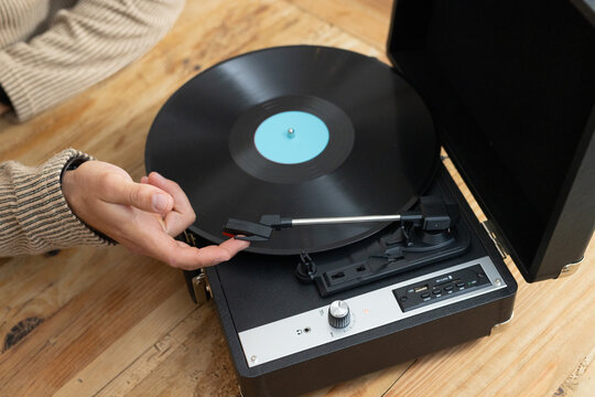 Man's left hand guiding tonearm to black vinyl on turntable on wood table in beige ribbed-sweater