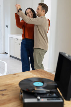Adult couple wearing rust sweater dancing while holding hands in kitchen by record player