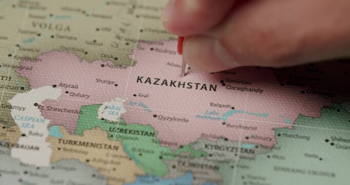 Close-up view of a person's hand placing a red flag pin on the country of Kazakhstan on a colorful political world map, marking a travel destination or a significant location
