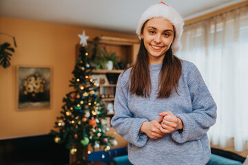 Naklejka premium Young woman wearing a Santa hat smiles while standing in a living room. A decorated Christmas tree and warm holiday atmosphere appear in the background.