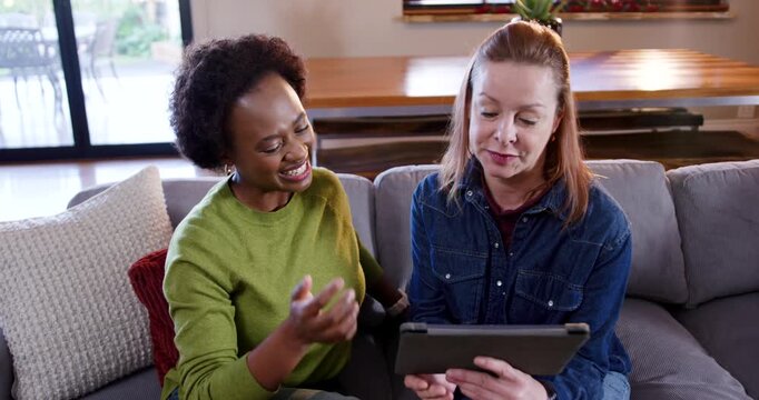 Diverse female friends on sofa, left greentop pointing tablet in case, denim pal handing to share