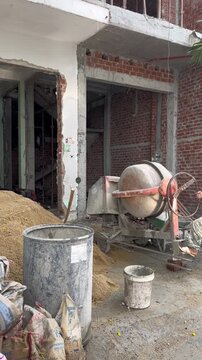 Construction site with a cement mixer, piles of sand, and bags of building materials. The area shows unfinished walls and exposed bricks.