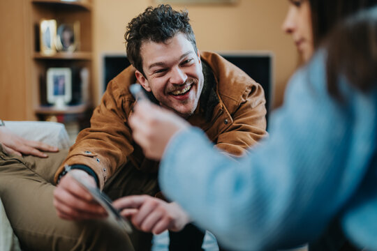 A smiling man and friend look at a smartphone while sharing photos in a cozy living room. Casual friends at home laugh and interact as they browse pictures together.