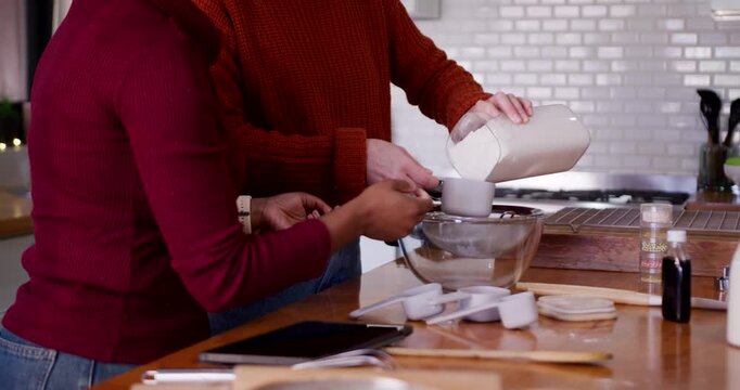 Diverse female pair measuring and sieving flour from jar at kitchen island using tablet for baking