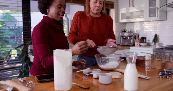 Diverse female friends sifting flour with sieve and measuring milk on kitchen island using tablet