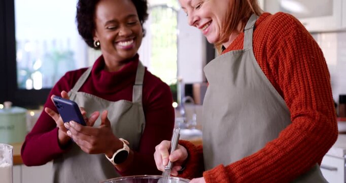 Diverse female cooks wearing aprons checking phone for recipe while whisking batter at wood counter