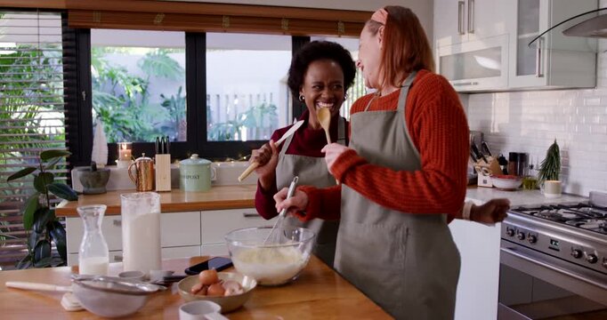 Diverse female friends preparing batter at wooden island, miming singing while whisking glass bowl