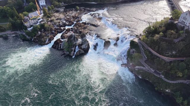 High aerial view of Rhine Falls in Switzerland, showing multiple cascades rushing around massive rock formations with cliffside viewing paths overlooking the turbulent water.